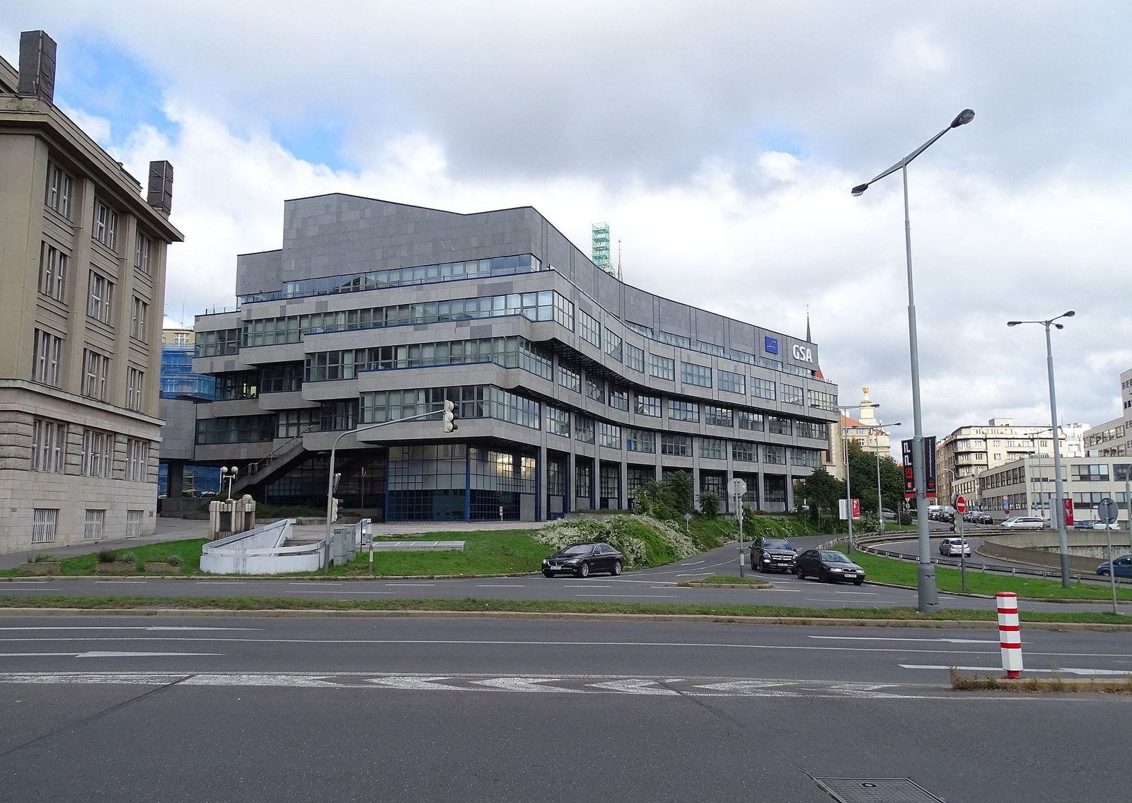 Prosklená terasovitá budova na rohu ulice, před ní silnice a tramvajová zastávka