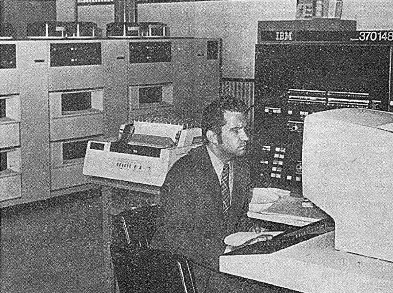 Muž v obleku sedí u konzole počítače IBM 370/148, vedle psací stroj konzole a řady skříní